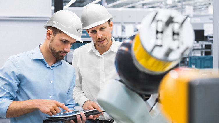 Two men with safety helmets in front of a machine looking at a circuit diagram